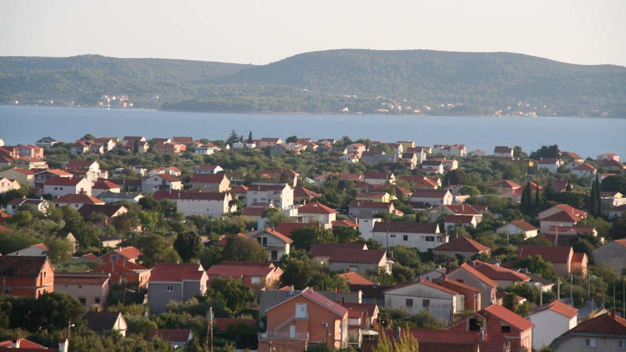 Kuće i manje stambene zgrade crvenih krovova u Sukošanu kraj Zadra, među borovima i zelenilom, s morem i otočnim brdima u pozadini, pri večernjem suncu